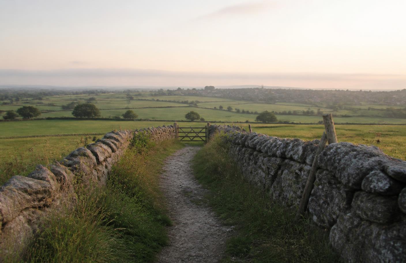 A stone-walled path leading through a wooden gate towards a distant English village